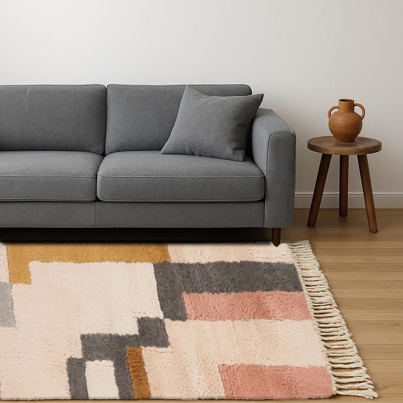 May include: A modern living room scene featuring a gray sofa with a matching pillow, a small wooden side table, and a patterned rug. The rug has a geometric design with blocks of cream, gray, pink, and gold. A terracotta vase sits on the table.