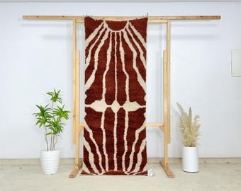 May include: A brown and cream-coloured rug with a sunburst design. The rug is displayed on a wooden frame, against a white background. The rug's pattern features vertical stripes radiating from a central point.