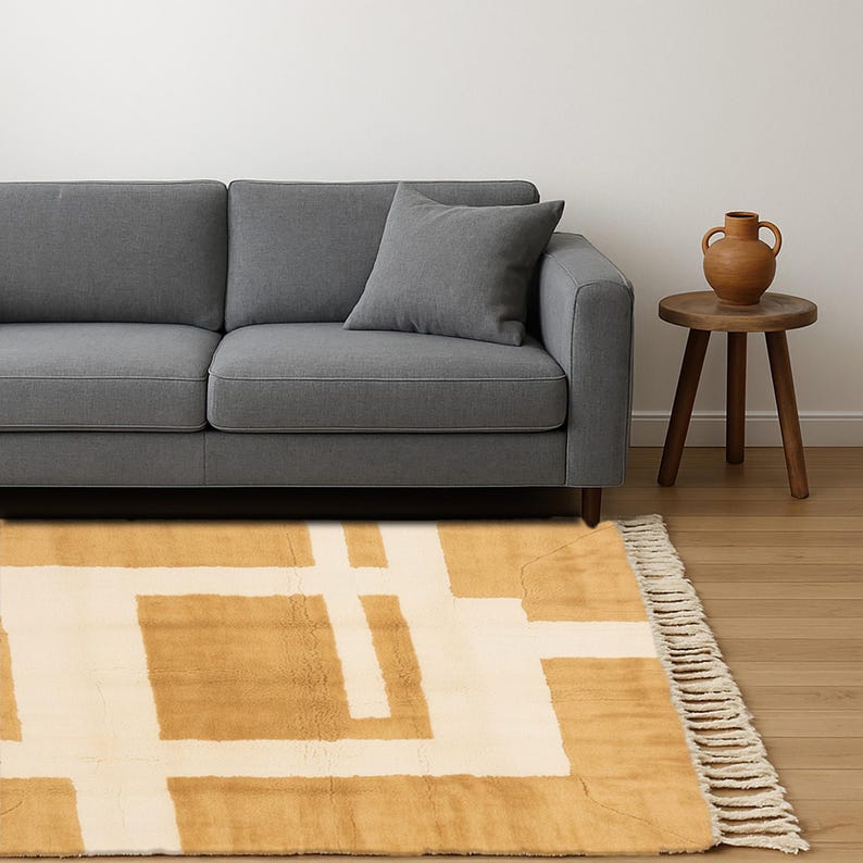 May include: A living room scene featuring a gray sofa with a matching pillow, a small wooden side table, and a terracotta vase. A mustard yellow and cream geometric patterned rug with fringe is in the foreground.
