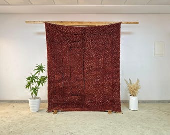 May include: A large, textured, burgundy-colored rug hangs from a wooden beam. The rug has a dense, nubby surface and is displayed against a white wall. A potted plant and dried flowers in a white vase are on either side.