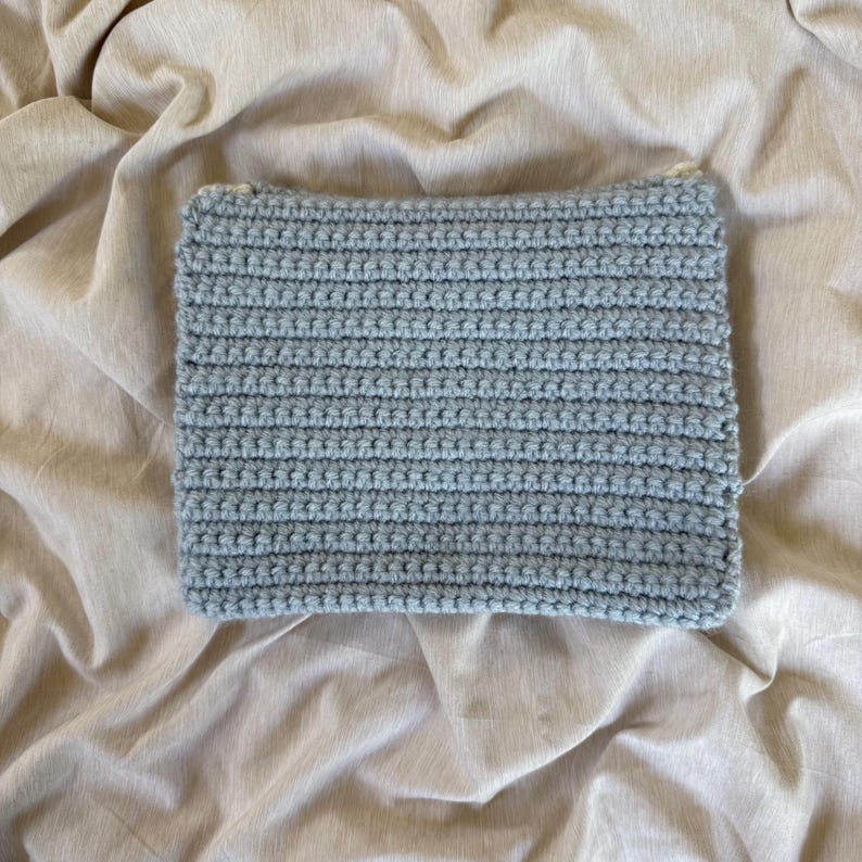 May include: A rectangular, light blue crochet item, possibly a pouch or small bag, is displayed on a neutral-toned, textured fabric. The crochet work features horizontal rows, creating a textured surface. The item's edges are neatly finished.