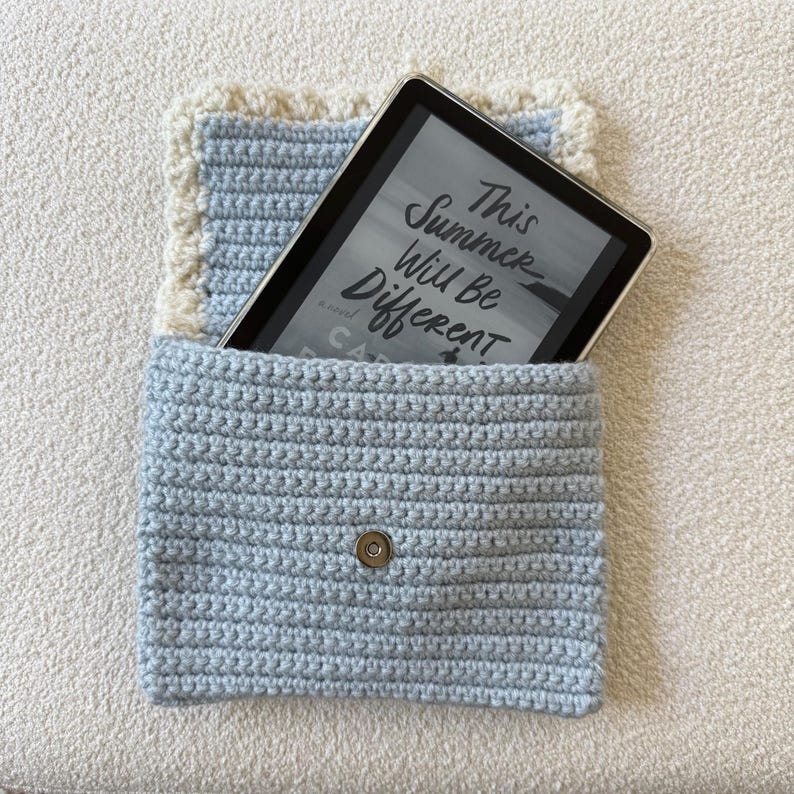 May include: A light blue crochet e-reader case with a cream-colored border. The case holds an e-reader displaying the text "This Summer Will Be Different". The case has a magnetic snap closure.