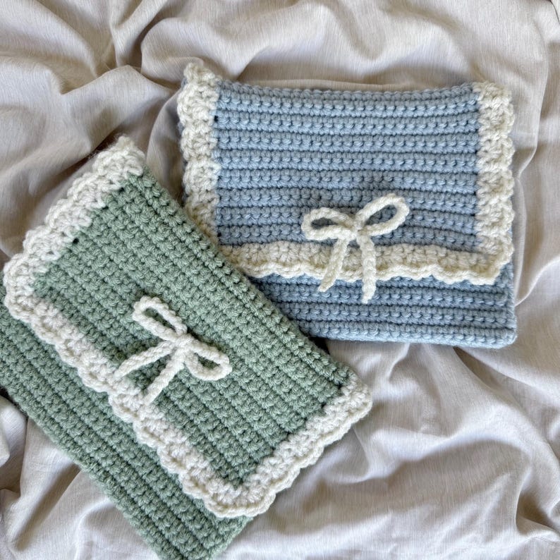 May include: Two crocheted pouches, one in a soft green and the other in light blue. Both have a white scalloped trim and a white bow closure. The pouches are handmade and appear to be soft and textured.