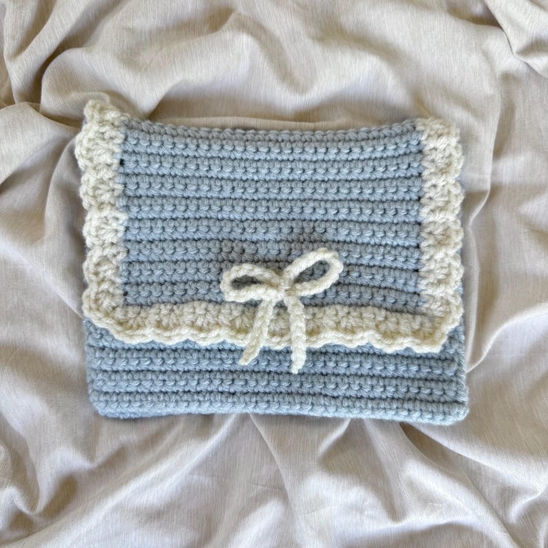 May include: A light blue crochet clutch with a cream-colored scalloped border and a cream bow closure. The handmade bag is rectangular and rests on a neutral-colored fabric background. The crochet stitches are visible.