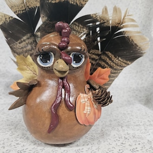 May include: A brown gourd turkey figurine featuring large blue eyes, a dark red wattle, and a fan of natural brown and black feathers. The turkey holds a small orange pumpkin cutout with a plaid ribbon and a brown pinecone in its wing.