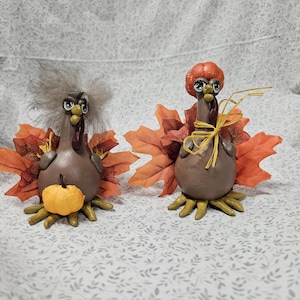 May include: Two ceramic turkey figurines with brown bodies, orange leaf accents, and whimsical details. One turkey has a fluffy gray head, the other a pumpkin head. Both have yellow feet and beaks, and are decorated for fall.