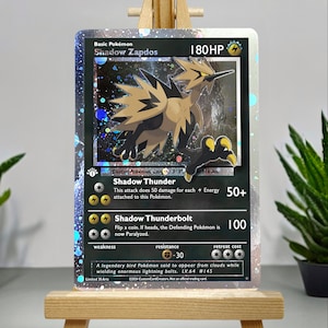 Può includere: Una carta da gioco di Pokemon olografica con Shadow Zapdos, un Pokemon di tipo elettrico con un design dorato e nero. La carta ha uno sfondo nero con un effetto olografico. Il testo della carta recita "Shadow Thunder" e "Shadow Thunderbolt".