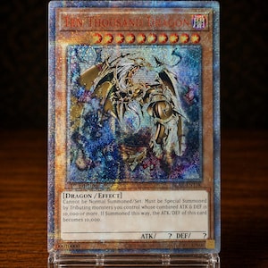 Peut inclure: Une carte à collectionner Ten Thousand Dragon avec une finition holographique aux tons bleus et dorés. La carte présente une illustration de dragon, le texte Ten Thousand Dragon et des règles de jeu dans un support en plastique transparent.