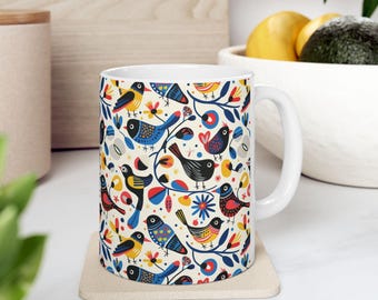 Puede incluir: Una taza de cerámica blanca con un patrón repetitivo de aves estilizadas en tonos azul, amarillo, rojo y negro, intercaladas con motivos florales y de hojas coloridos. La taza tiene un asa lisa y una forma cilíndrica con interior blanco.