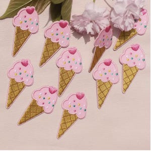 Op de afbeelding: Een verzameling van negen geborduurde ijsje-patches. Elke patch heeft een lichtroze bolletje ijs met een klein roze hartje en gekleurde spikkels, geplaatst op een goudkleurige wafelhoorn met een ruitvormig textuurpatroon.