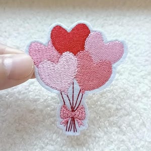 May include: Embroidered patch showing a bouquet of heart-shaped balloons in red and pink hues. The balloons are tied with a pink bow and strings. The patch has a white border.
