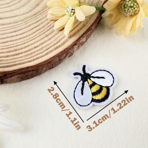 Pode incluir: Um patch de abelha bordado com contorno branco, antenas pretas e corpo listrado amarelo e preto. O patch mede 2,8 cm por 3,1 cm. Em um fundo claro, com uma fatia de madeira e flores amarelas.