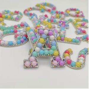 May include: A collection of beaded letters in various pastel colours, including pink, yellow, blue, and green. The letters are outlined in silver and decorated with small rhinestones. The letters are scattered on a white surface.