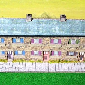 May include: A scale model of a row of stone houses featuring a blue-grey shingled roof, three chimneys, and two levels of windows with blue, purple, and green shutters. The structure includes three front doors and a grey tiled pavement at the base.