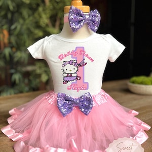 May include: A white short-sleeved bodysuit featuring a Hello Kitty graphic, the text "Birthday Princess" and "Kaylee", and a large number one. The outfit includes a pink tulle tutu with a purple sequin bow and a matching purple sequin headband.