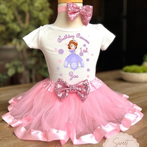 May include: A white short-sleeved bodysuit with a graphic of a princess and the text "Birthday Princess 1st Zoe". The outfit includes a pink tutu skirt, a sequined bow, and a matching headband with a sequined bow.