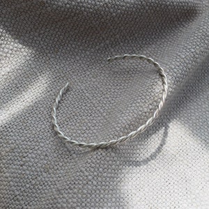 May include: A silver-coloured, open cuff bracelet with a twisted rope design. The bracelet is resting on a textured, grey fabric. The bracelet's simple design makes it a versatile accessory.