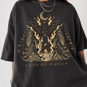 May include: A charcoal gray oversized t-shirt featuring a gold-toned graphic print of a winged figure, a crescent moon, and floral elements. The text "girls will be girls" is printed in gold at the bottom of the central illustration.