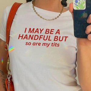 May include: A white short-sleeved t-shirt featuring the red text I MAY BE A HANDFUL BUT so are my tits printed on the front. The shirt is worn with a gold chain necklace and an orange shoulder bag with a silver clasp and a small charm.