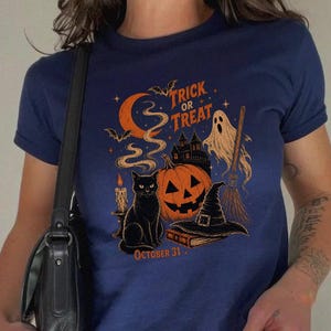 Op de afbeelding: Een marineblauw t-shirt met korte mouwen en een grafische print met de tekst "TRICK OR TREAT" en "OCTOBER 31". Het ontwerp bevat een oranje maansikkel, een zwarte kat, een pompoen, een spook, een heksenhoed en een bezemsteel.