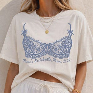May include: An ivory short-sleeved t-shirt featuring a blue graphic of a lace bralette with bows on the straps. Below the graphic, the text reads "Olivia's Bachelorette, Miami 2026" in a cursive script. The shirt has a crew neckline and a relaxed fit.