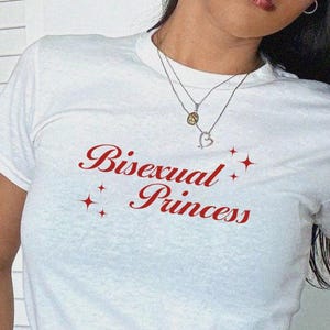 Könnte beinhalten: Weißes T-Shirt mit dem Text "Bisexual Princess" in roter Schrift und Sternen. Das Shirt hat einen Rundhalsausschnitt.