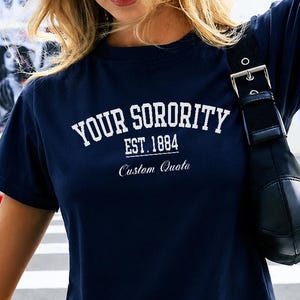 May include: A navy blue short-sleeved crop top featuring white text that reads YOUR SORORITY EST. 1884 and Custom Quote in a script font. The shirt is paired with light gray sweatpants with a white drawstring and a black shoulder bag.