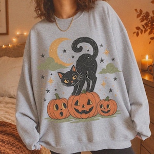 Può includere: Una felpa girocollo grigio chiaro con una stampa grafica raffigurante un gatto nero in piedi su una zucca di Halloween. Il disegno include due zucche più piccole, una luna crescente gialla, nuvole verdi e stelle nere sparse.