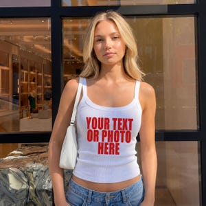 May include: A white ribbed tank top featuring the red text YOUR TEXT OR PHOTO HERE printed on the front. The garment has a scoop neckline and thick shoulder straps, paired with a small white shoulder bag and blue denim jeans.