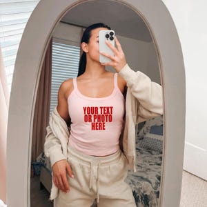 May include: A light pink tank top featuring the red text "YOUR TEXT OR PHOTO HERE" printed on the chest, worn with a beige zip-up hoodie and matching beige sweatpants. The garment is shown in a mirror reflection with a smartphone held in front.