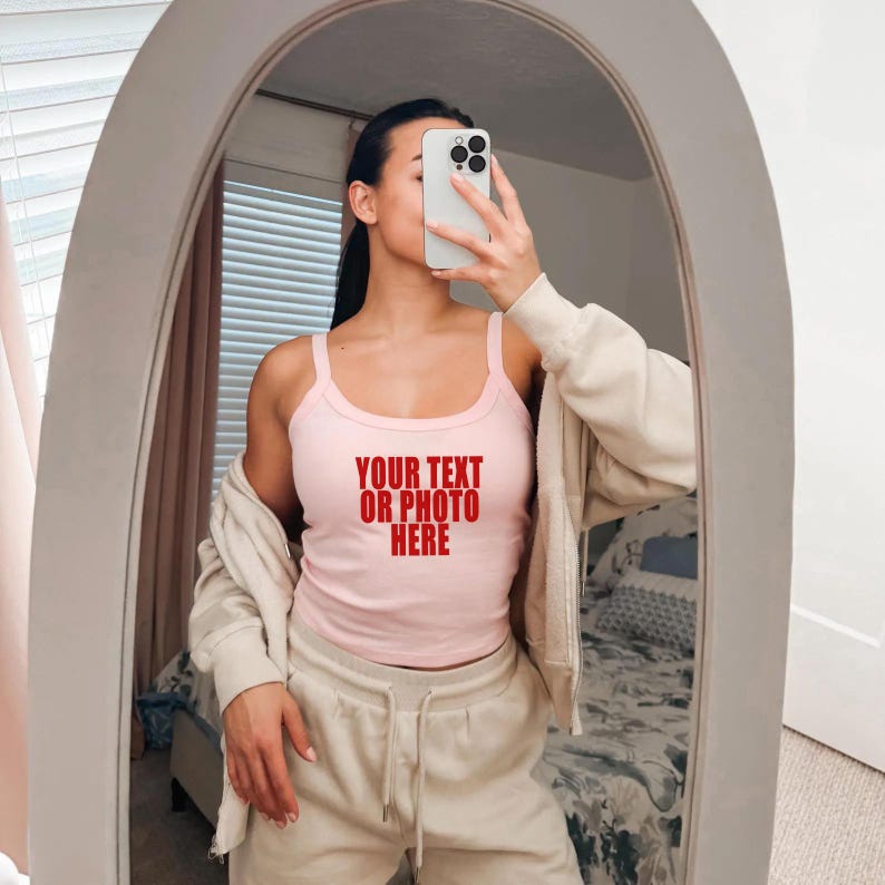 May include: A light pink tank top featuring the red text "YOUR TEXT OR PHOTO HERE" printed on the chest, worn with a beige zip-up hoodie and matching beige sweatpants. The garment is shown in a mirror reflection with a smartphone held in front.