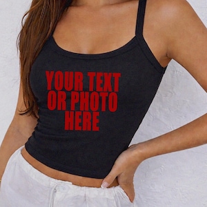 May include: A black ribbed tank top featuring a scoop neckline and thick shoulder straps. The center of the garment displays the red text "YOUR TEXT OR PHOTO HERE" in a bold, sans-serif font, indicating a customizable area for personal designs.