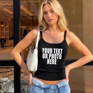 May include: A black ribbed tank top featuring the white text YOUR TEXT OR PHOTO HERE printed on the front, paired with a white shoulder bag and light blue denim jeans. The tank top has a scoop neckline and wide shoulder straps.