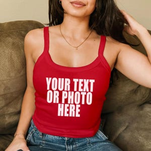 May include: A bright red sleeveless tank top featuring a scoop neckline and thick shoulder straps. The center of the chest displays the white text "YOUR TEXT OR PHOTO HERE" in a bold, sans-serif font, indicating a customizable garment design.