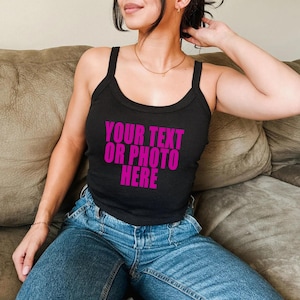 May include: A black sleeveless tank top with a scoop neckline and thick shoulder straps. The front of the garment features the text "YOUR TEXT OR PHOTO HERE" printed in a bold, bright magenta pink color across the center of the chest area.