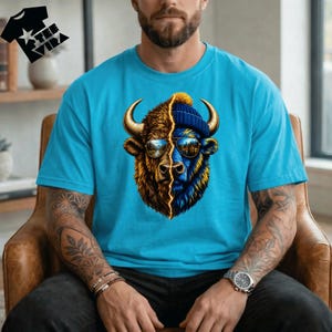 Peut inclure: T-shirt bleu turquoise avec un motif de bison divisé en deux. Une moitié est marron et or, l'autre bleue et jaune, avec des lunettes de soleil reflétant un paysage. Le bison porte un bonnet bleu. Le t-shirt porte le texte "TEE VIRX".
