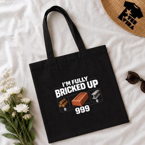 Peut inclure: Sac fourre-tout noir avec le texte "I'M FULLY BRICKED UP" et des graphiques de bois, une brique et de l'acier. Le graphique de la brique porte le numéro 999 en dessous. Le sac a deux anses noires.