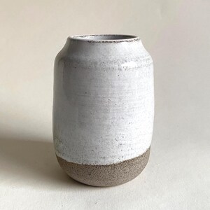 Könnte beinhalten: Eine handgefertigte Keramikvase mit zylindrischer Form. Der obere Teil ist weiß, während der Boden aus strukturiertem, erdigem Braun besteht. Die Vase hat eine leicht ausgestellte Öffnung und eignet sich zur Präsentation von Blumen oder als dekoratives Element.