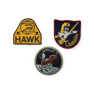 Pode incluir: Três patches bordados com temas militares e espaciais. Um patch amarelo apresenta um falcão e a palavra "HAWK". Outro patch mostra um jato e a palavra "Crusader". O terceiro patch retrata uma águia pousando na lua com o texto "Apollo 11".