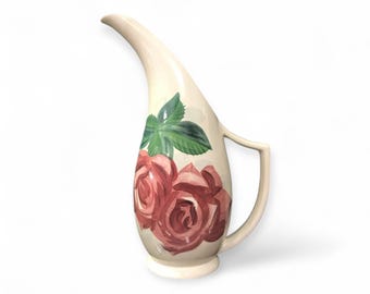 May include: A white ceramic pitcher with a curved spout and a handle. The pitcher is decorated with a hand-painted design of pink roses and green leaves.