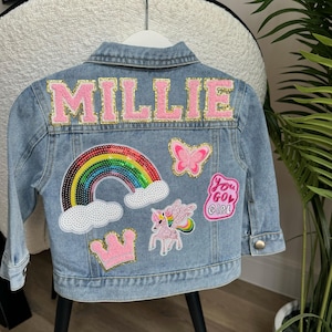 May include: Pale blue denim jacket with the name "MILLIE" in pink letters with gold trim on the back. The jacket has patches of a rainbow, butterfly, unicorn, crown, and the text "You Go Girl".