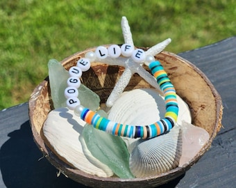 Puede incluir: Un brazalete de cuentas blancas con las palabras "Pogue Life" escritas en letras negras. El brazalete está en un cuenco de cáscara de coco con conchas marinas y una estrella de mar.