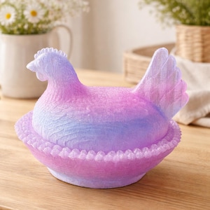 Puede incluir: Un recipiente decorativo con forma de gallina en tonos rosa, morado y azul. La gallina se asienta sobre una base a juego con un borde festoneado. El recipiente está sobre una superficie de madera, con flores en el fondo.