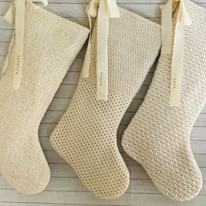 Puede incluir: Tres calcetines de punto de color crema con nombres personalizados, "Natalie", "Tracy" y "Zack", colgados en un fondo de madera blanca.