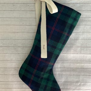 May include: A Christmas stocking featuring a dark green and navy blue plaid pattern with thin red accent lines. The stocking is adorned with a cream-colored fabric ribbon tied in a bow at the top, with the word Dad printed on one ribbon tail.