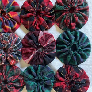 May include: A selection of fabric circles with a tartan design. The circles are in shades of red, green, and blue, with yellow accents. The fabric is gathered in the centre, creating a textured, circular shape. These could be used for crafting or as decorative elements.