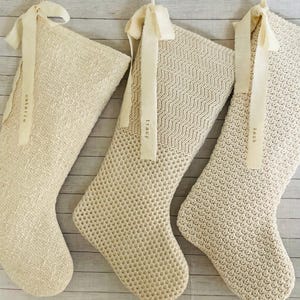 May include: Three cream-colored knit Christmas stockings hanging vertically. Each stocking features a unique textured weave pattern and a cream ribbon tied at the top. The ribbons are printed with the names natalie, tracy, and zack respectively.