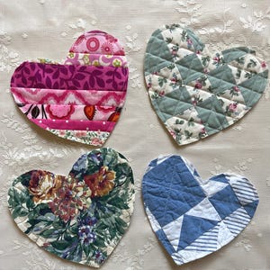 May include: Four heart-shaped quilted coasters in various fabric patterns. One coaster features pink and purple stripes, another has a floral design on a green background, and a third has a blue and white geometric pattern. The fourth coaster has a floral design.