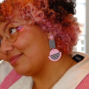 May include: Pink and black geometric earrings with a checkered pattern and a half-circle design. The earrings are worn by a person with pink hair.