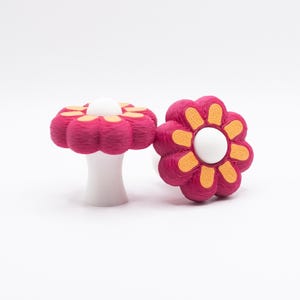 Op de afbeelding: Twee bloemvormige kastknoppen in magenta met oranje en gele details. Elke knop heeft een wit midden. Eén staat rechtop op een witte basis, de andere ligt plat. Geschikt voor een kinderkamer.